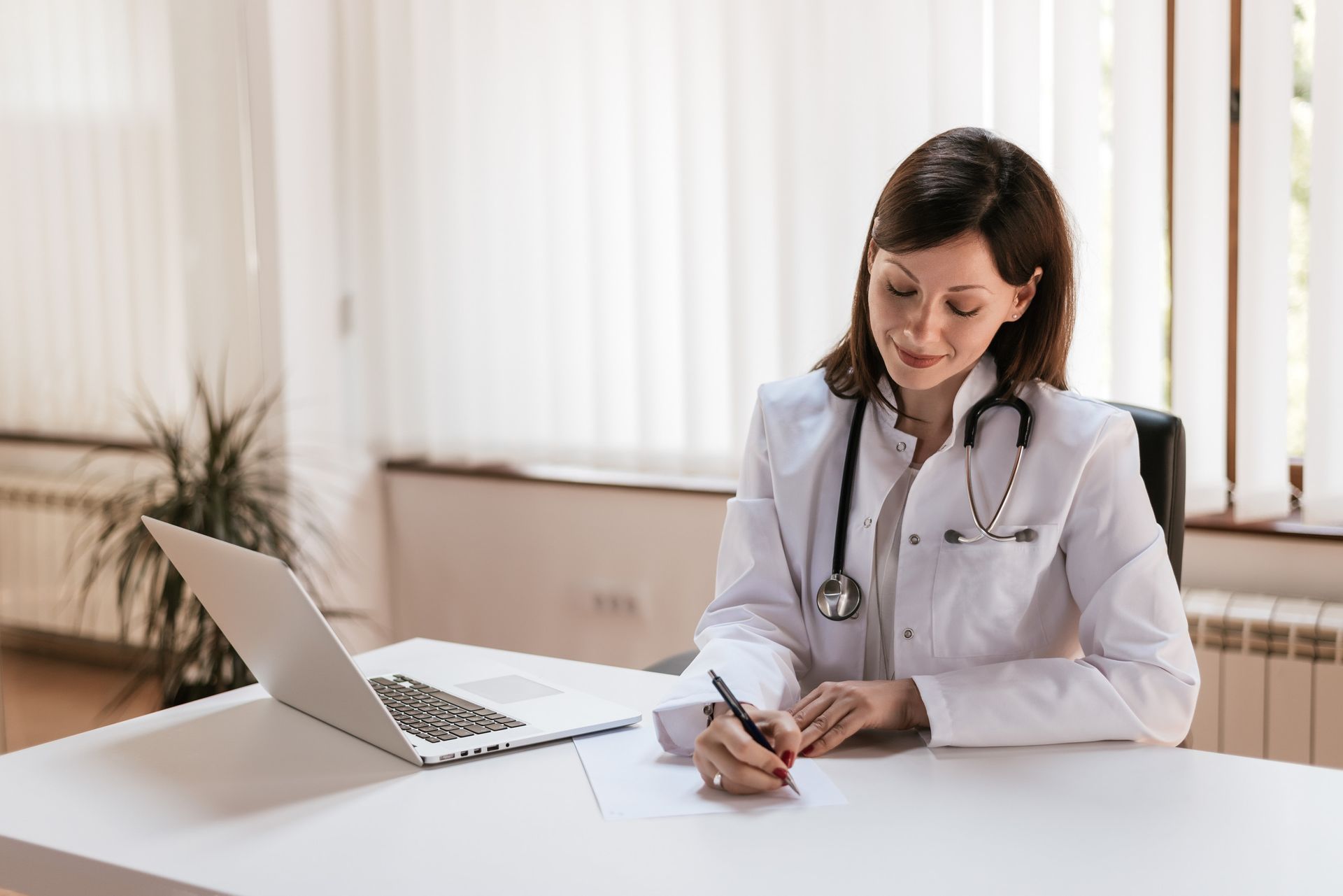 Una médica está sentada en un escritorio con una computadora portátil y escribiendo en una hoja de papel.