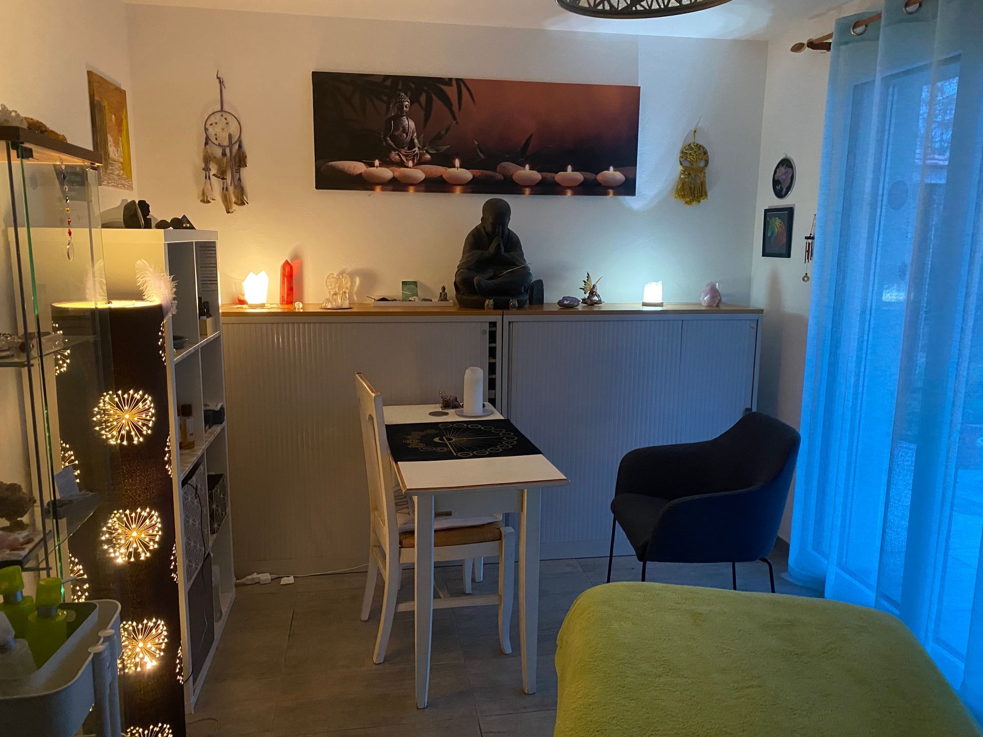 Chambre confortable avec statue de Bouddha, bougies, petite table, fauteuil et étagères éclairées.