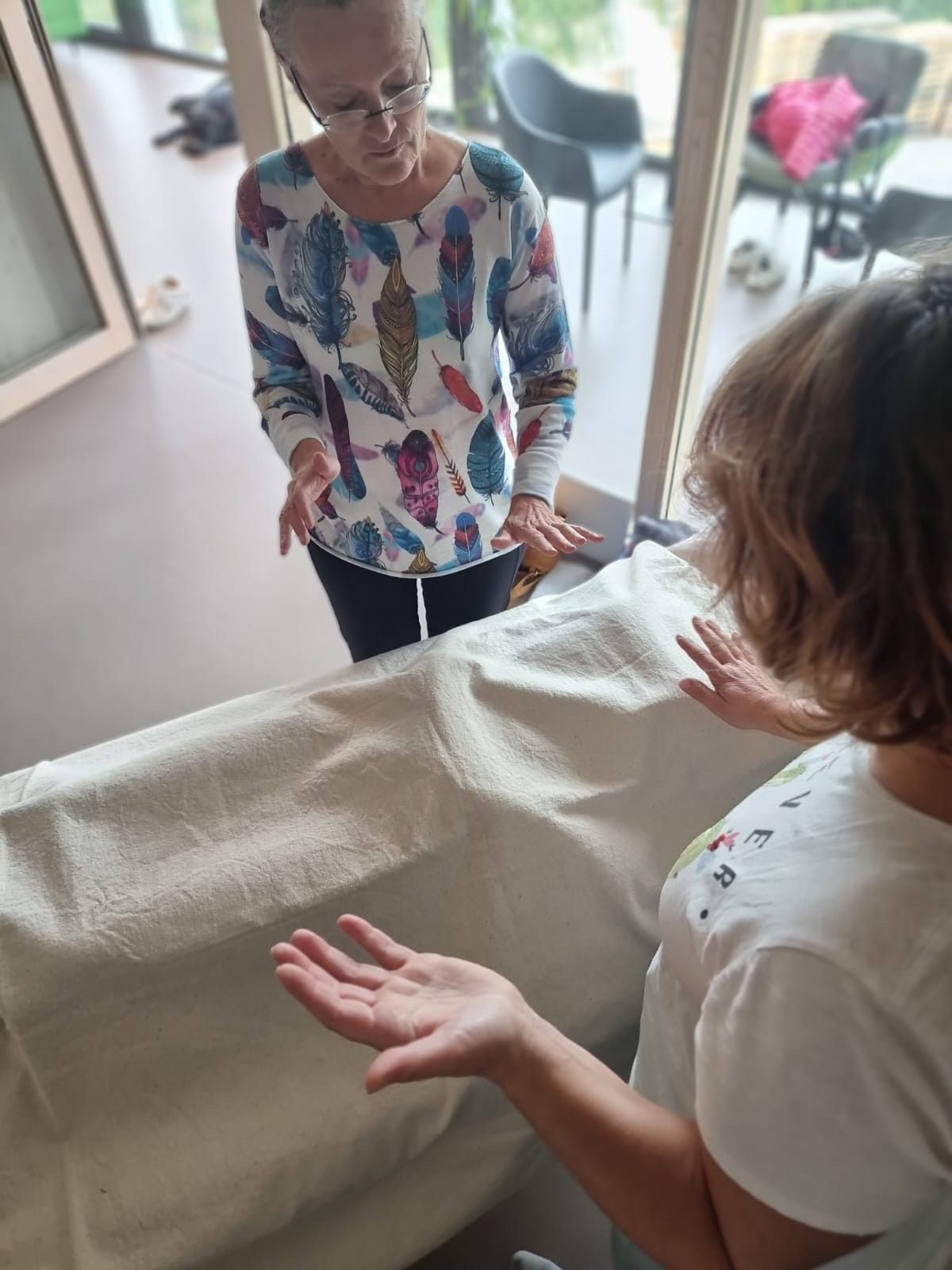 Deux personnes interagissent, l'une d'elles faisant un geste au-dessus d'un drapé. Intérieur avec de grandes fenêtres.