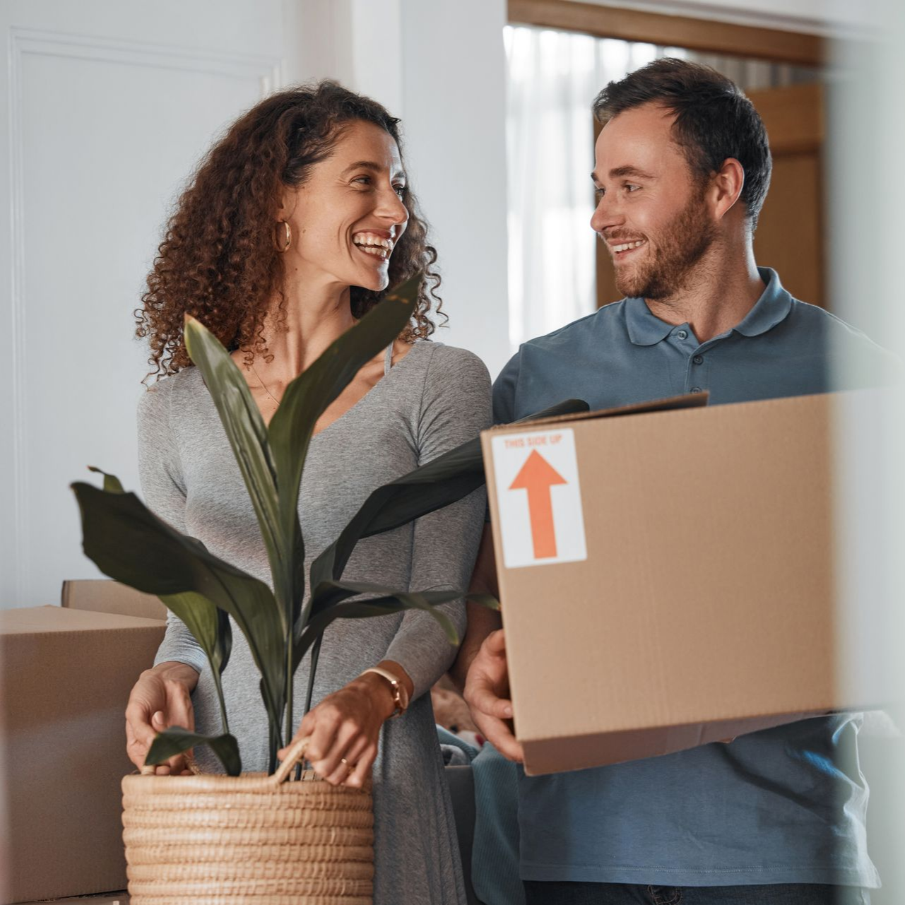 Un couple sourit en déménageant des cartons et une plante dans leur nouvelle maison.