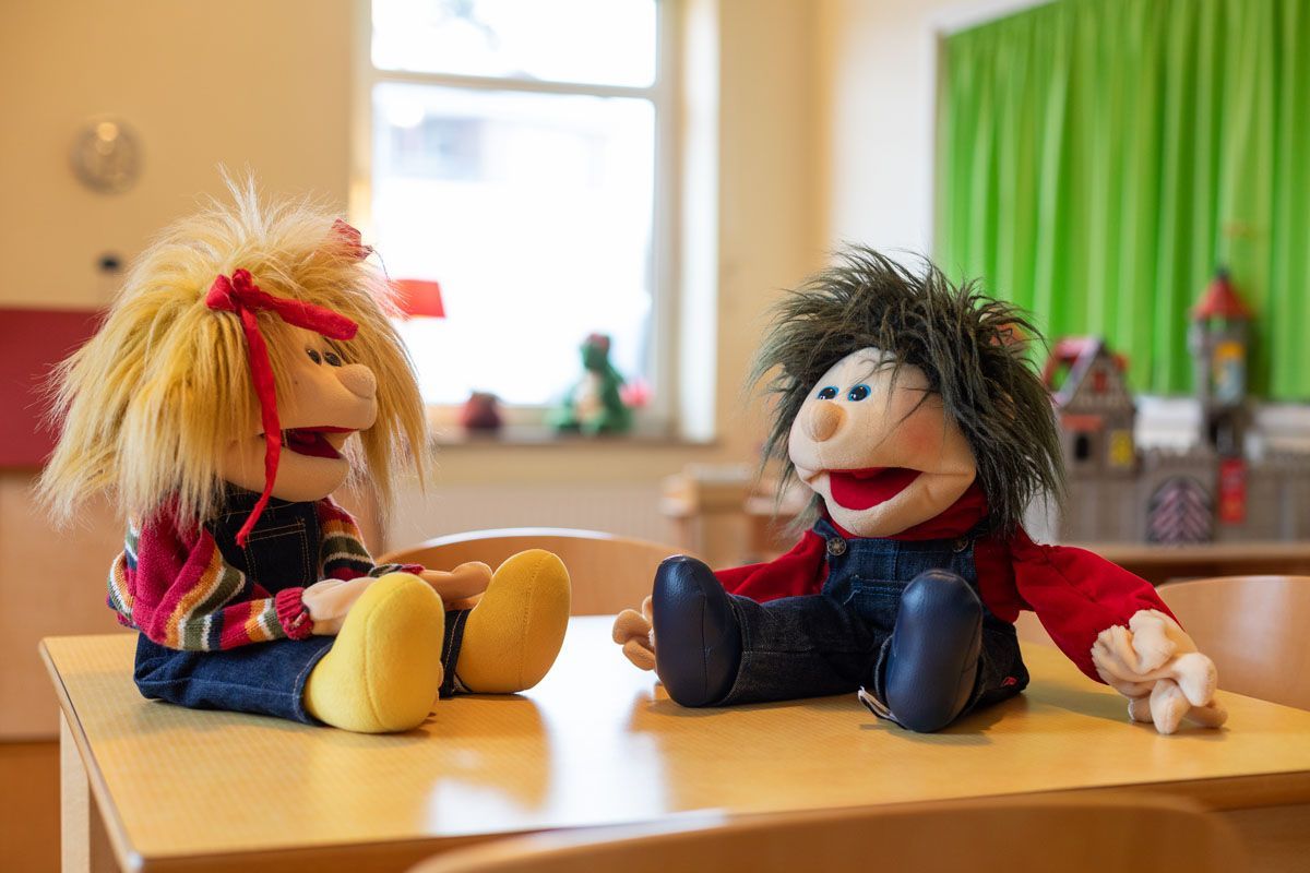 Zwei Stoffpuppen mit wildem Haar und Jeanslatzhosen sitzen einander auf einem kleinen Tisch in einem hellen Klassenzimmer gegenüber.