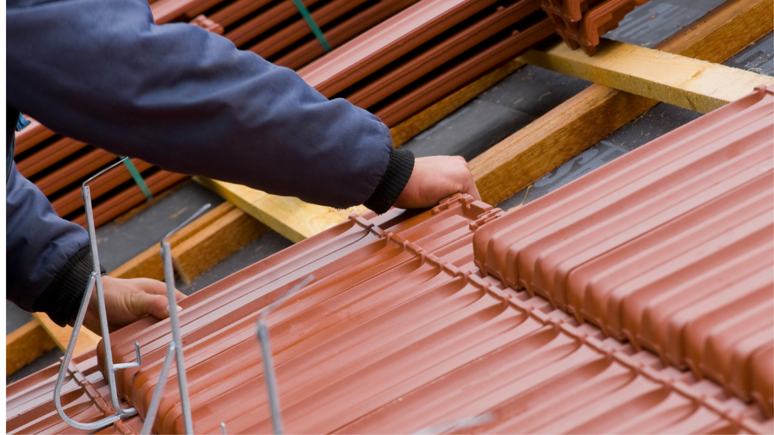 Un ouvrier installe des tuiles rouges sur une charpente en bois, sur le toit d'un bâtiment.