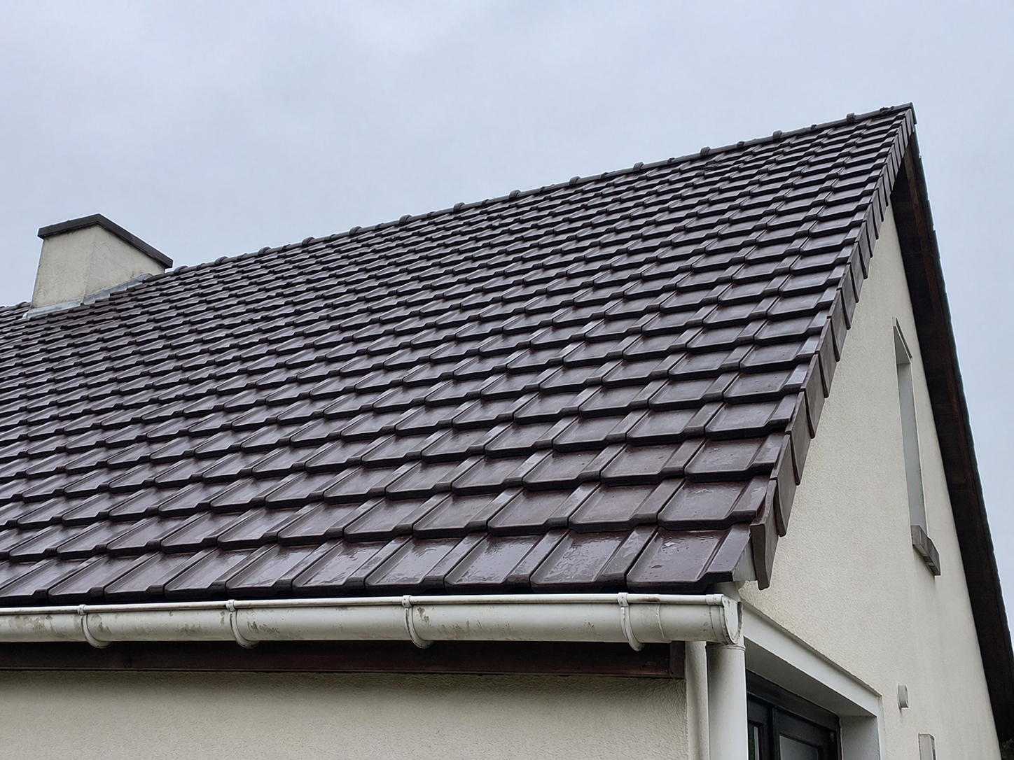 Vue en contre-plongée d'un toit de tuiles brun foncé sur une maison beige, se détachant sur un ciel nuageux.