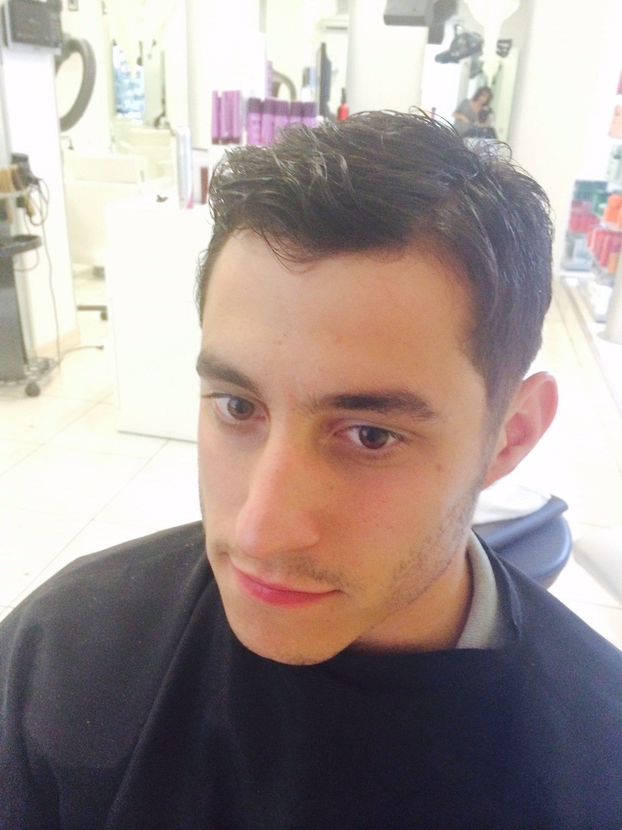 Mann mit dunklem Haar in einem Salon, trägt einen schwarzen Umhang und blickt mit neutralem Gesichtsausdruck nach unten.