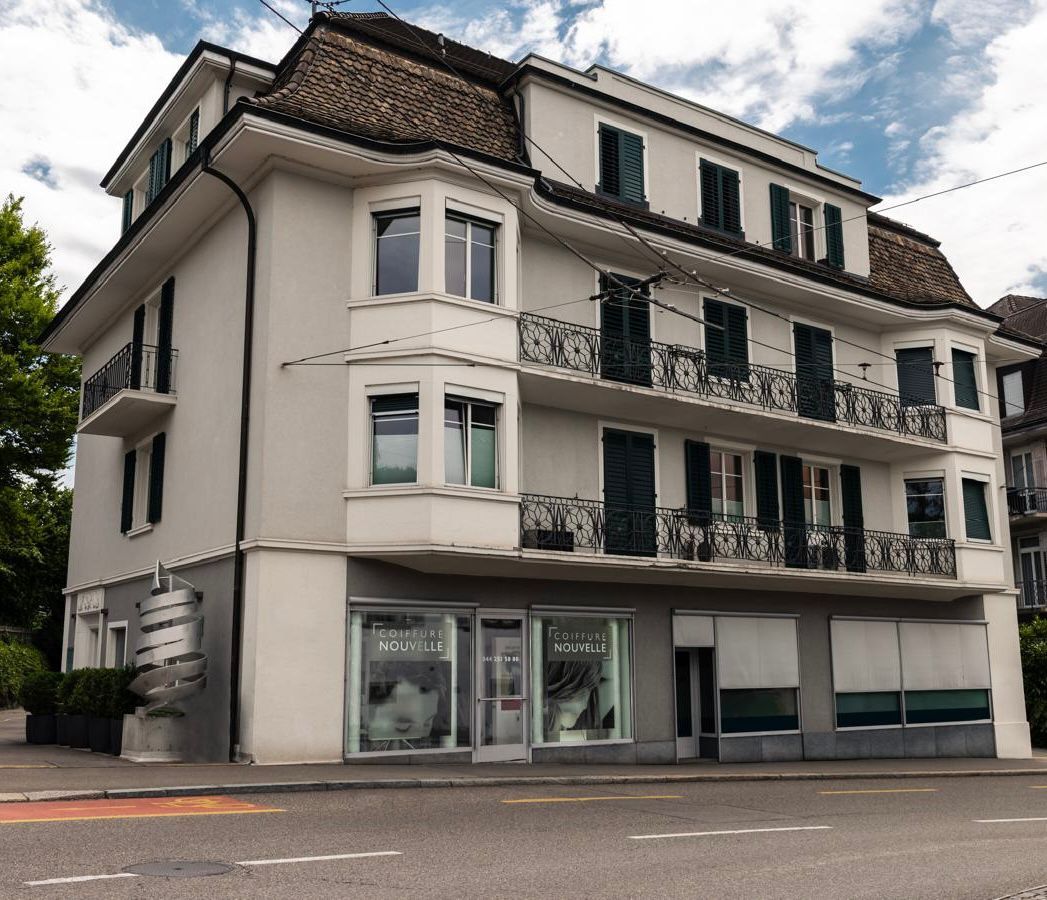 Dreistöckiges Gebäude mit Geschäften im Erdgeschoss; hellgraue Fassade, dunkle Fensterläden und braunes Dach.