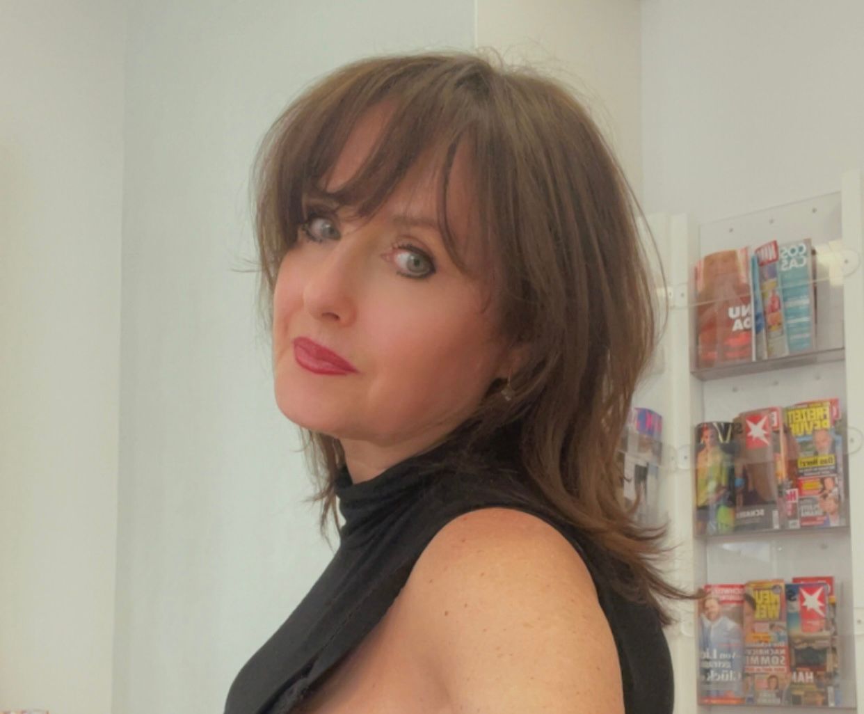 Frau mit braunem Haar und rotem Lippenstift, die vor Zeitschriften über ihre Schulter blickt.
