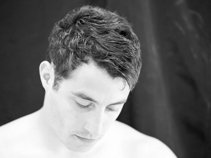 Mann mit nach unten gerichtetem Blick, ohne Hemd, dunklem, welligem Haar vor schwarzem Hintergrund.