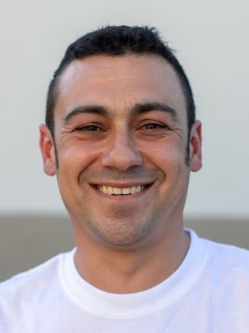 Mann mit kurzen dunklen Haaren, lächelnd, trägt ein weisses Hemd, vor hellem Hintergrund.