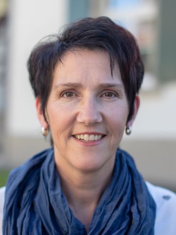 Frau mit kurzen dunklen Haaren, die einen blauen Schal und eine weisse Jacke trägt, lächelt im Freien.