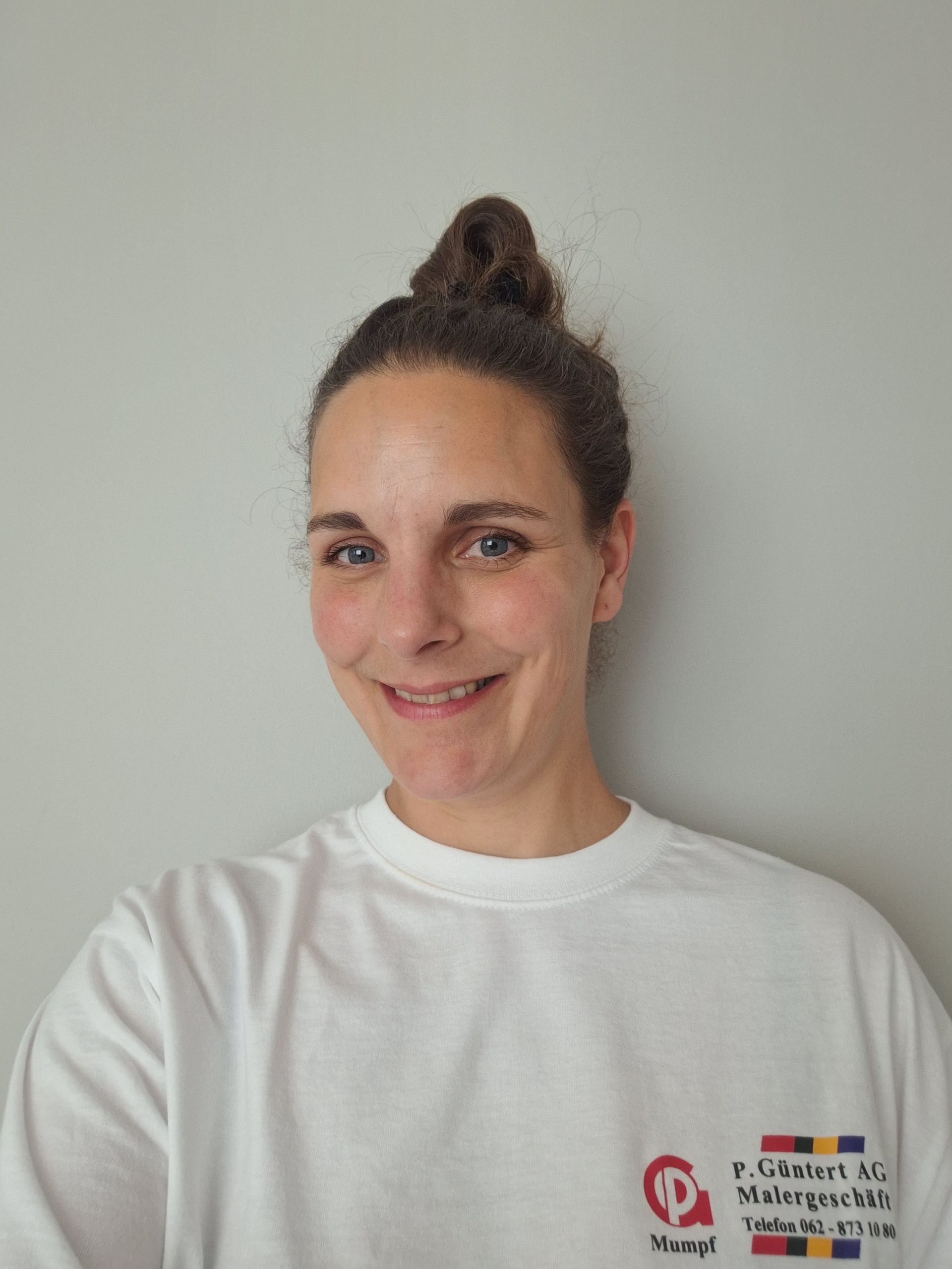 Frau mit braunem, zum Dutt gebundenem Haar lächelt und trägt ein weisses Hemd mit Logo auf dem Hemd.