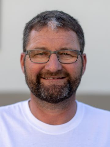 Ein Mann mit Brille und Bart lächelt im Freien. Er trägt ein weisses T-Shirt.