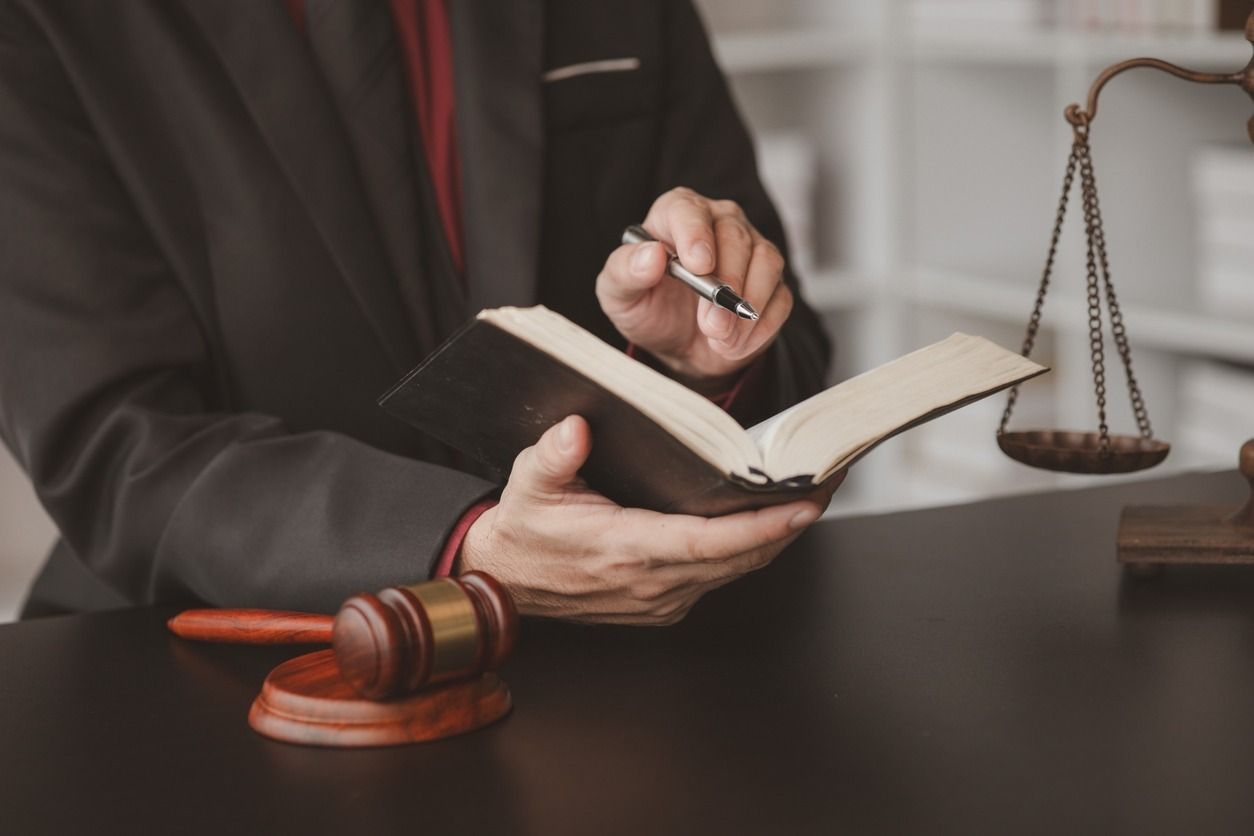 Primer plano de una persona con traje sosteniendo un libro y un bolígrafo, junto a un mazo y una balanza de la justicia sobre un escritorio.