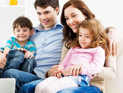 Una familia de cuatro personas, sonriendo mientras mira una computadora portátil en un sofá; incluye una madre, un padre y dos niños pequeños.