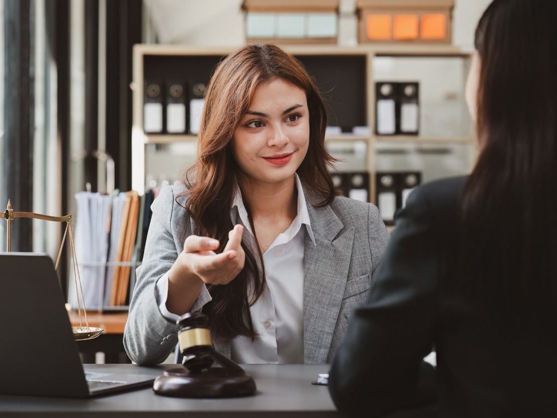Dos mujeres de traje en una oficina legal, una gesticulando y hablando, la otra frente a ella, con un mazo y una computadora portátil sobre el escritorio.