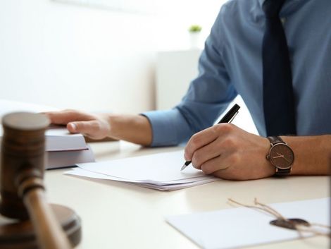 Un hombre con una camisa azul escribe en un papel en un escritorio, al lado de un mazo y documentos.