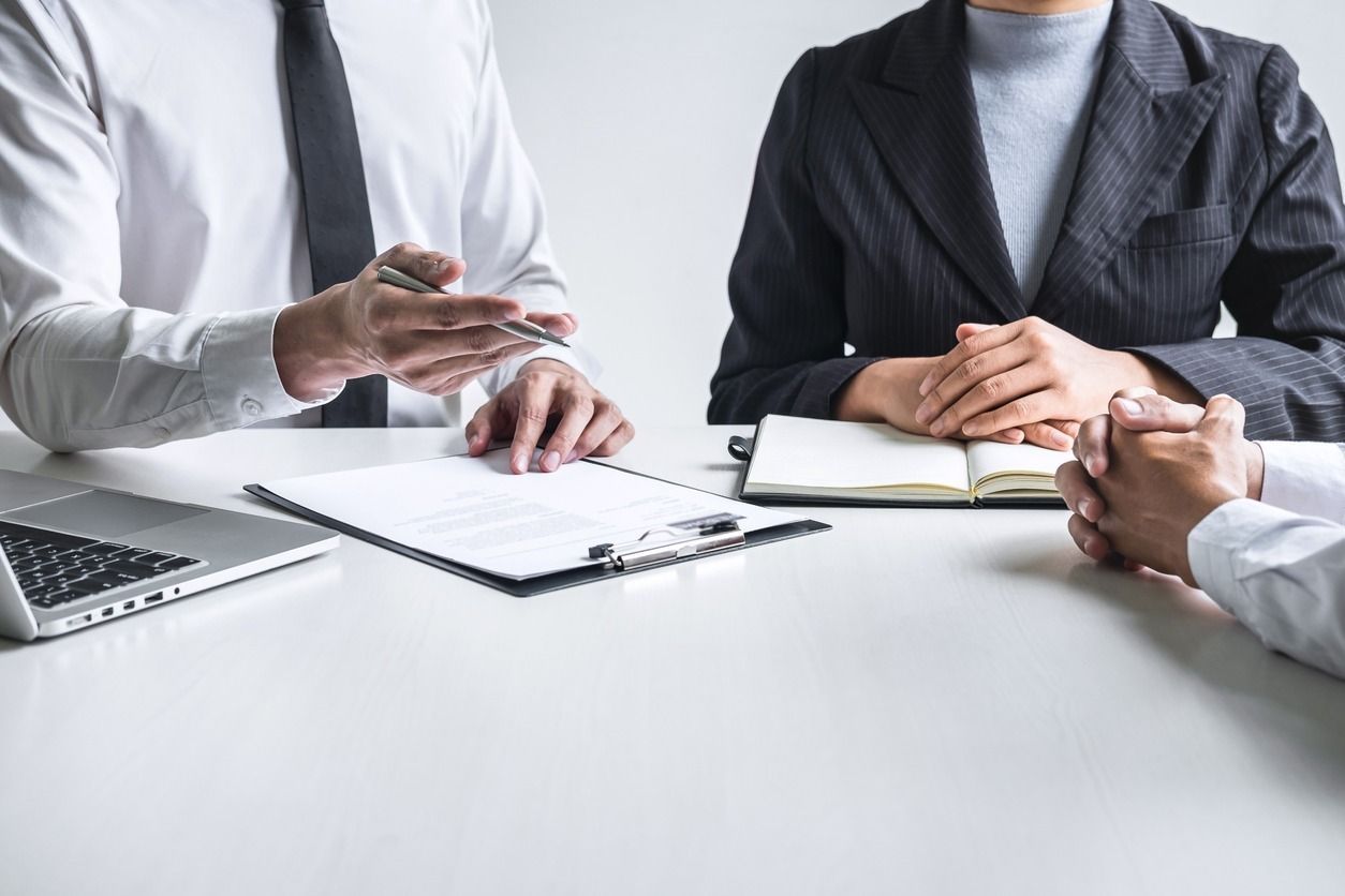 Empresarios sentados a la mesa, uno de ellos señalando documentos. Dos llevan traje y uno lleva camisa blanca. Portátil, bloc de notas y portapapeles sobre la mesa.