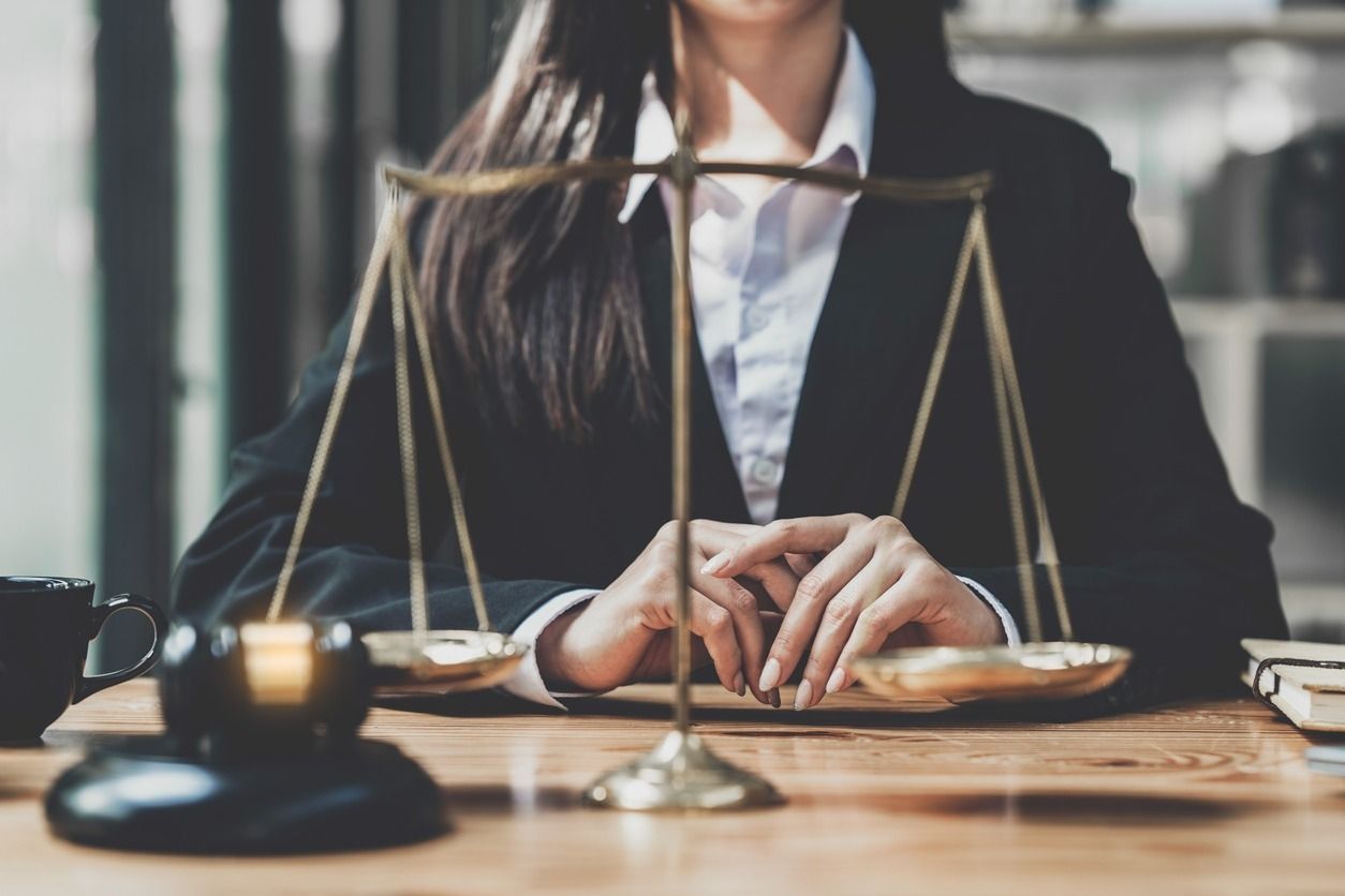 Mujer con un blazer detrás de una balanza dorada sobre un escritorio de madera, con las manos entrelazadas.