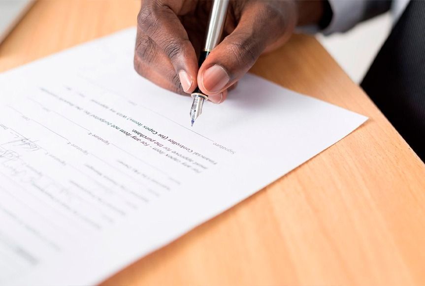 Primer plano de una mano negra firmando un documento con un bolígrafo sobre un escritorio de madera.