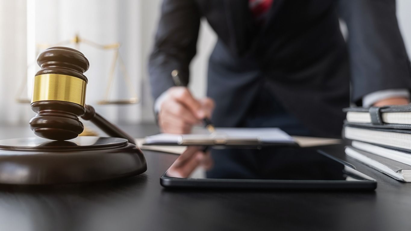 Un mazo y una balanza sobre un escritorio con una persona con traje escribiendo en el fondo, probablemente en un tribunal.