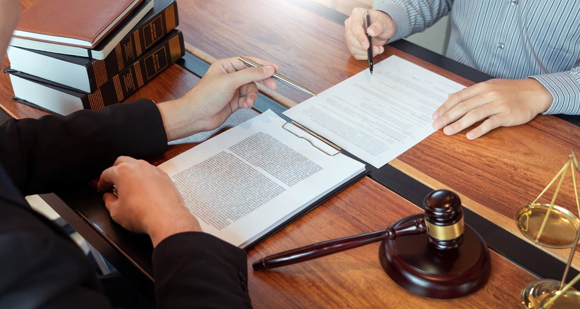 Dos personas en un escritorio, una firmando documentos, con libros de leyes, un mazo y una balanza de la justicia en el fondo.