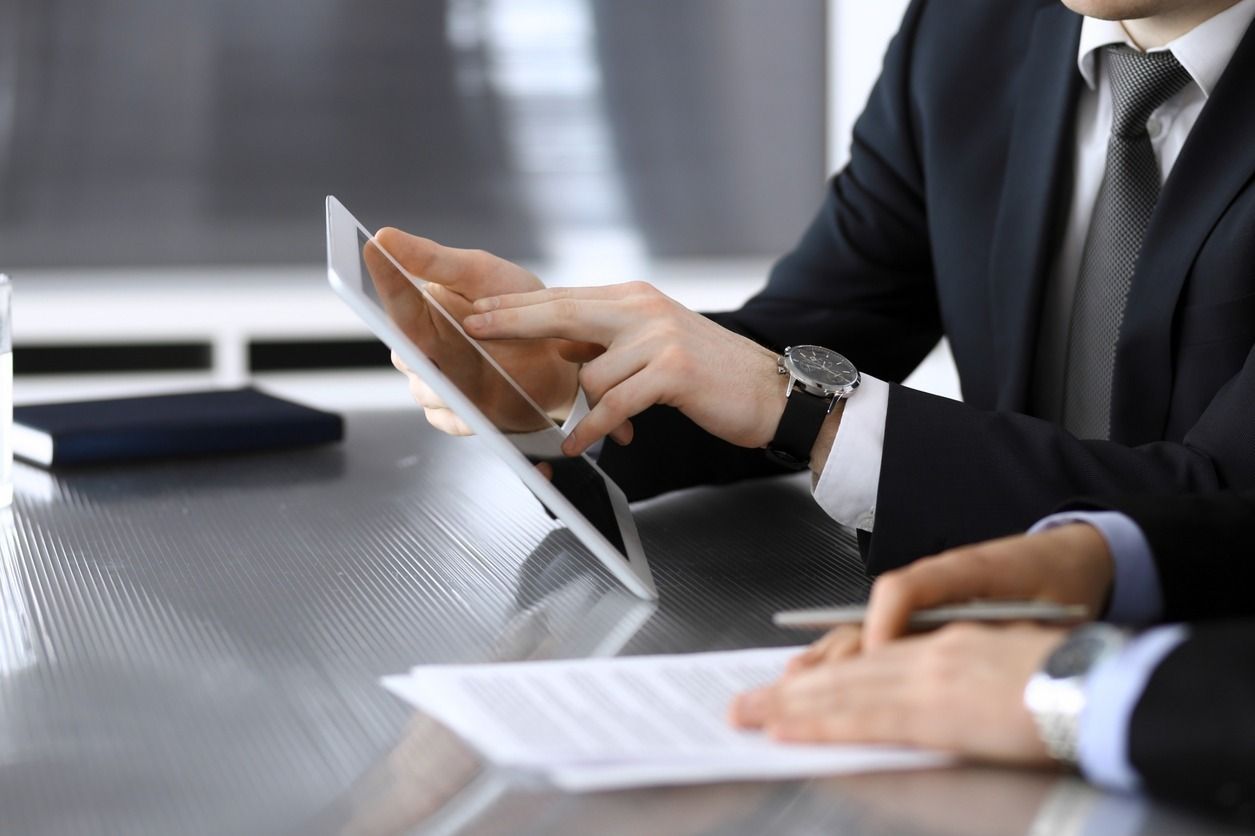 Dos hombres de negocios, con traje, revisan documentos y una tableta en una mesa. Uno de ellos señala la pantalla.