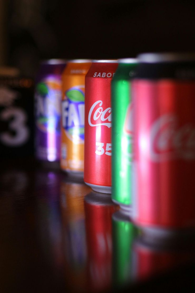Hilera de latas de refresco de colores sobre una superficie reflectante, con una lata roja de Coca-Cola en primer plano.