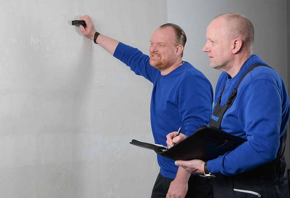 Zwei Männer in blauen Hemden vermessen eine Wand; einer hält ein Klemmbrett, der andere benutzt ein Werkzeug.