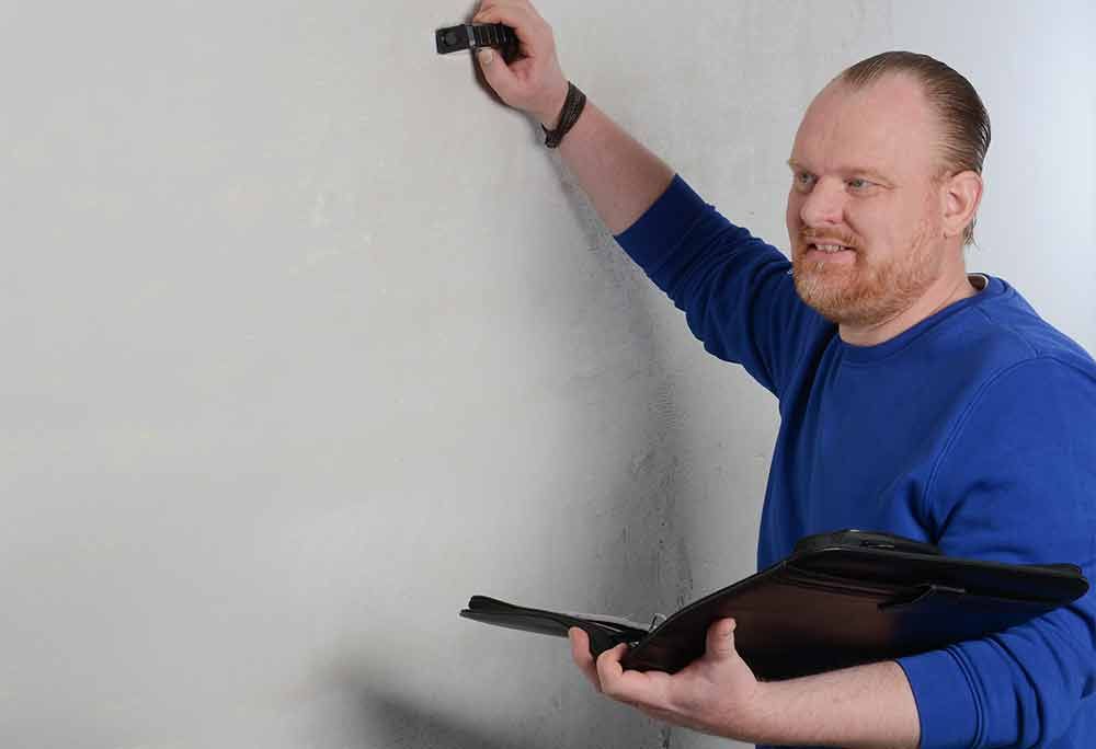 Mann im blauen Hemd benutzt ein Mobilgerät und einen Laptop vor einer weißen Wand