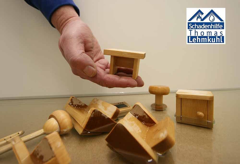 Eine Hand hält ein kleines Holzspielzeug; im Hintergrund liegen verstreute Holzklötze auf einem Tisch; im Hintergrund ist ein Logo zu sehen.
