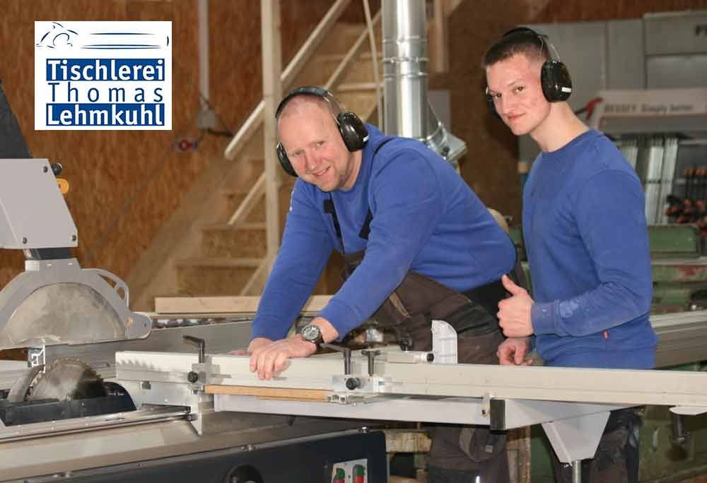 Zwei Männer bedienen eine Tischkreissäge in einer Holzwerkstatt; sie tragen blaue Oberteile und Headsets.