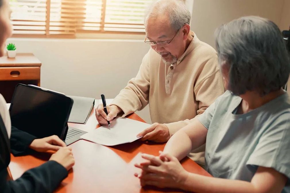 Un anciano firma documentos con una mujer; otra mujer observa sentada en una mesa con una computadora portátil.