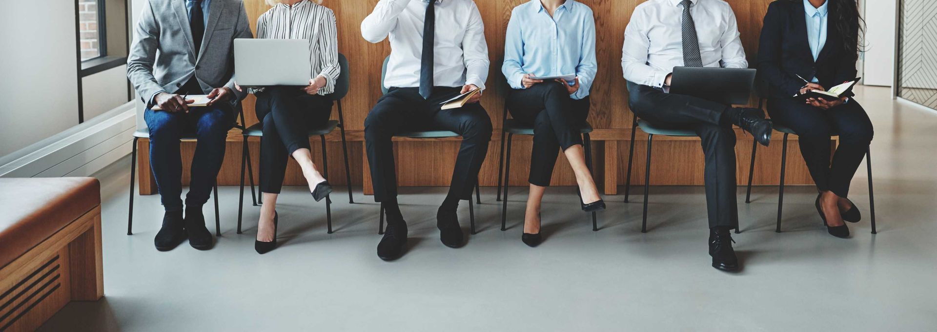 Personas vestidas de negocios sentadas en sillas, probablemente esperando entrevistas en una oficina.