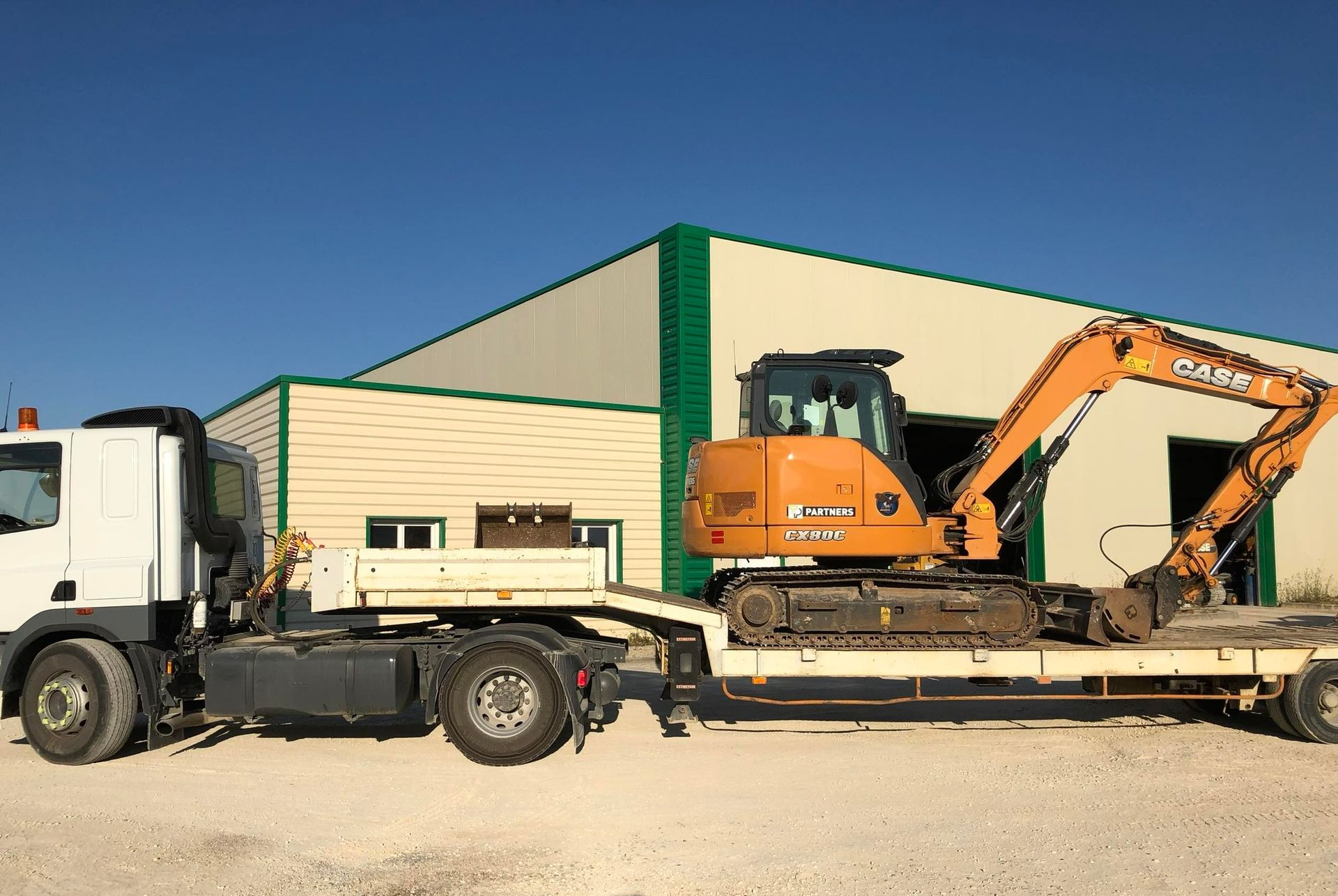 Un camion de chantier transportant une excavatrice orange à côté d'un bâtiment industriel beige.