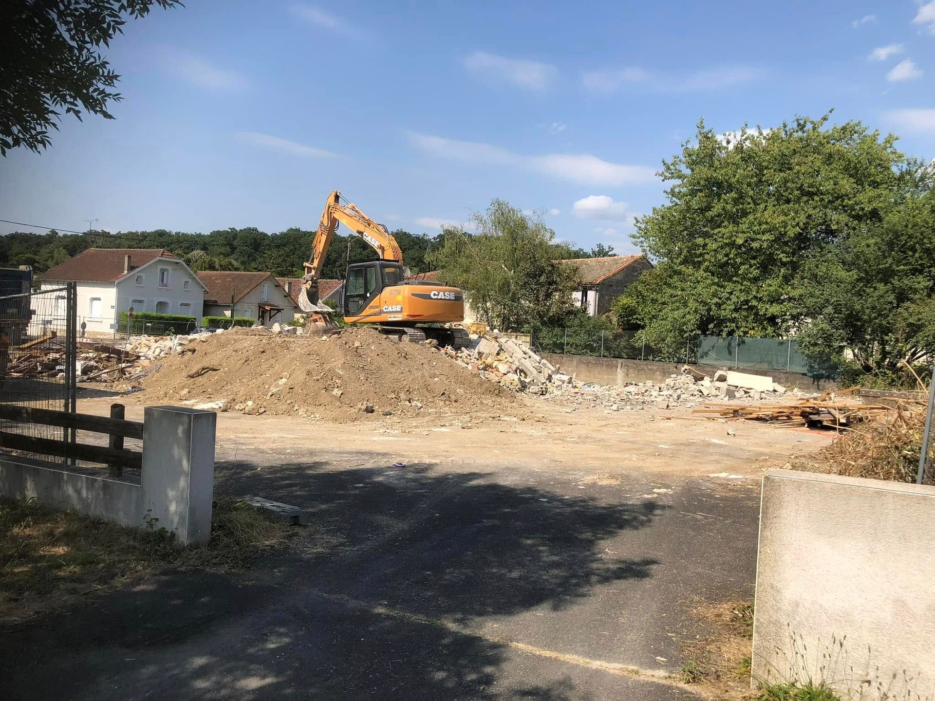 Une pelleteuse sur un chantier de construction en terre battue, avec des débris et des maisons en arrière-plan.