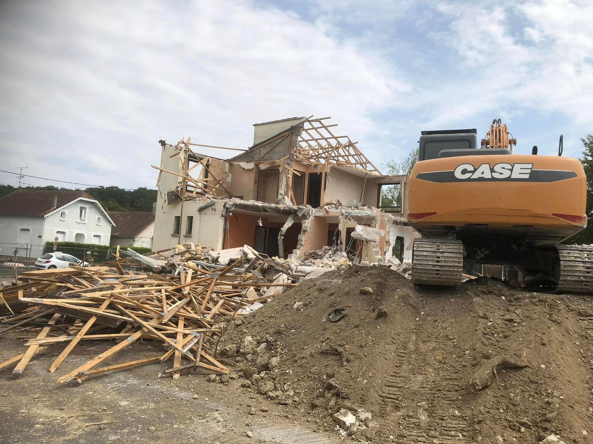 Une pelleteuse jaune se dresse devant une maison à moitié démolie, avec des tas de débris de bois au premier plan.