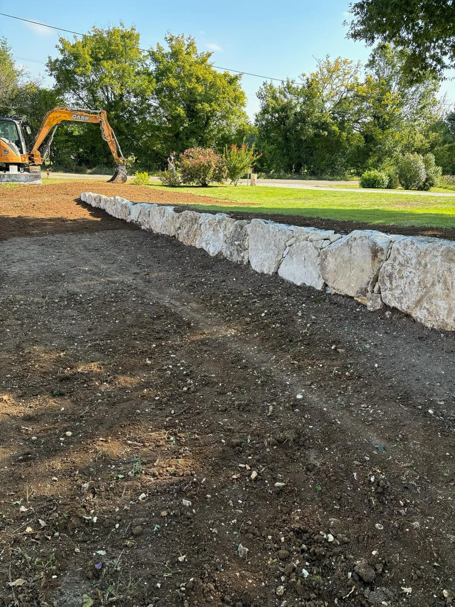 Un mur de soutènement en pierre construit dans un paysage en pente, avec des engins de chantier visibles en arrière-plan.