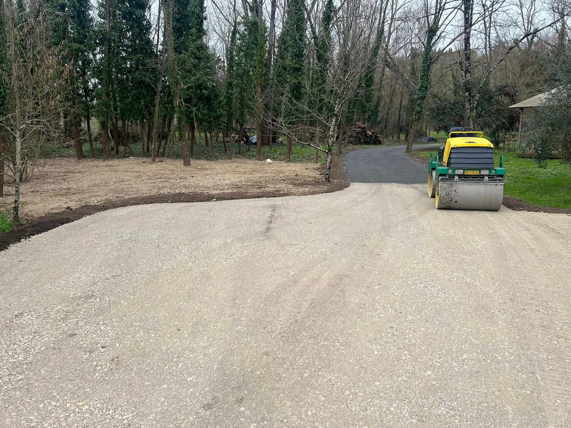 Un rouleau compresseur jaune est posé sur une allée de gravier, à côté d'un chemin pavé, dans un cadre boisé et extérieur.