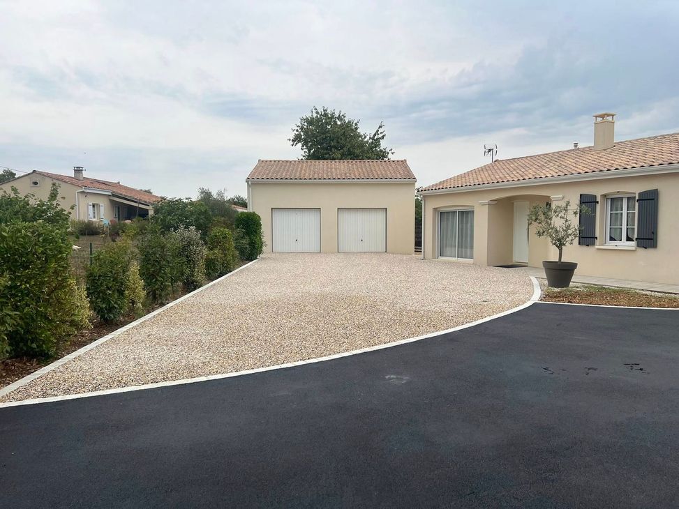 Une maison individuelle beige avec garage, une allée en gravier clair bordée d'asphalte et d'une haie verte.