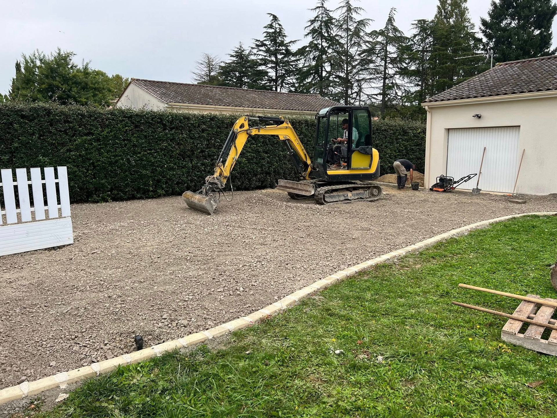 Une pelleteuse sur une surface gravillonnée près d'une haie et d'un garage blanc, une personne travaillant à proximité.