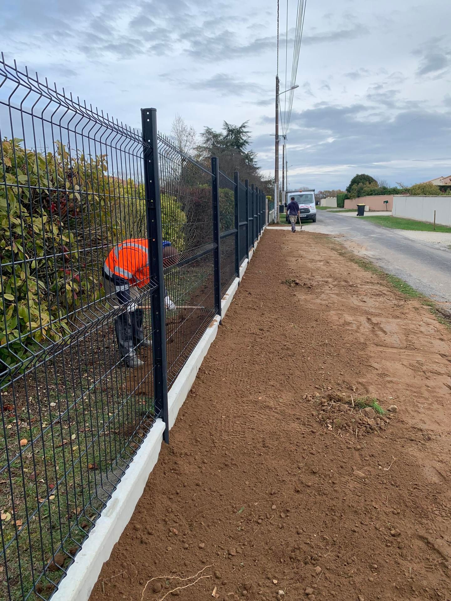 Deux ouvriers installent une clôture métallique noire avec une base en béton le long d'un chemin de terre.