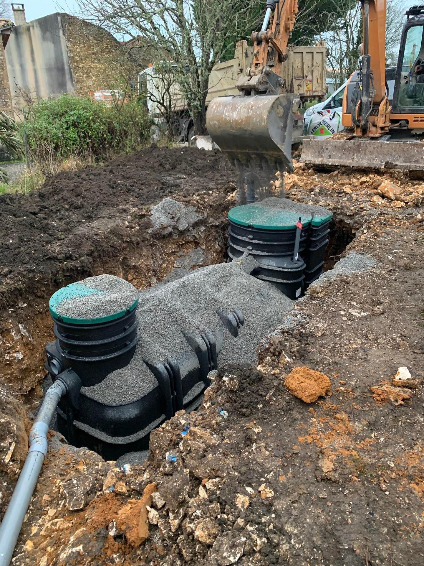 Une excavatrice installe deux fosses septiques en plastique noir dans une tranchée remplie de gravier.