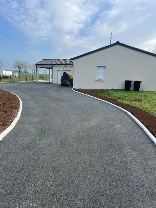 Une allée asphaltée neuve, bordée de bordures en béton blanc, mène à une maison moderne aux tons clairs.
