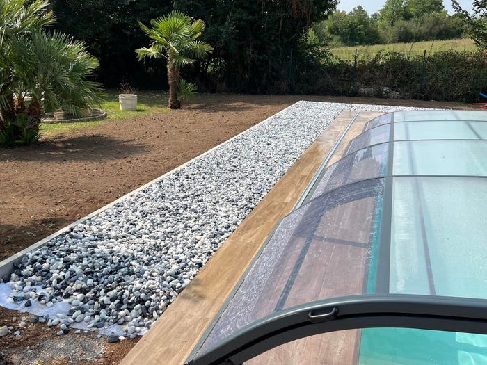 Structure à toit de verre jouxtant une bande de gravier et un jardin paysager avec arbres et haie.