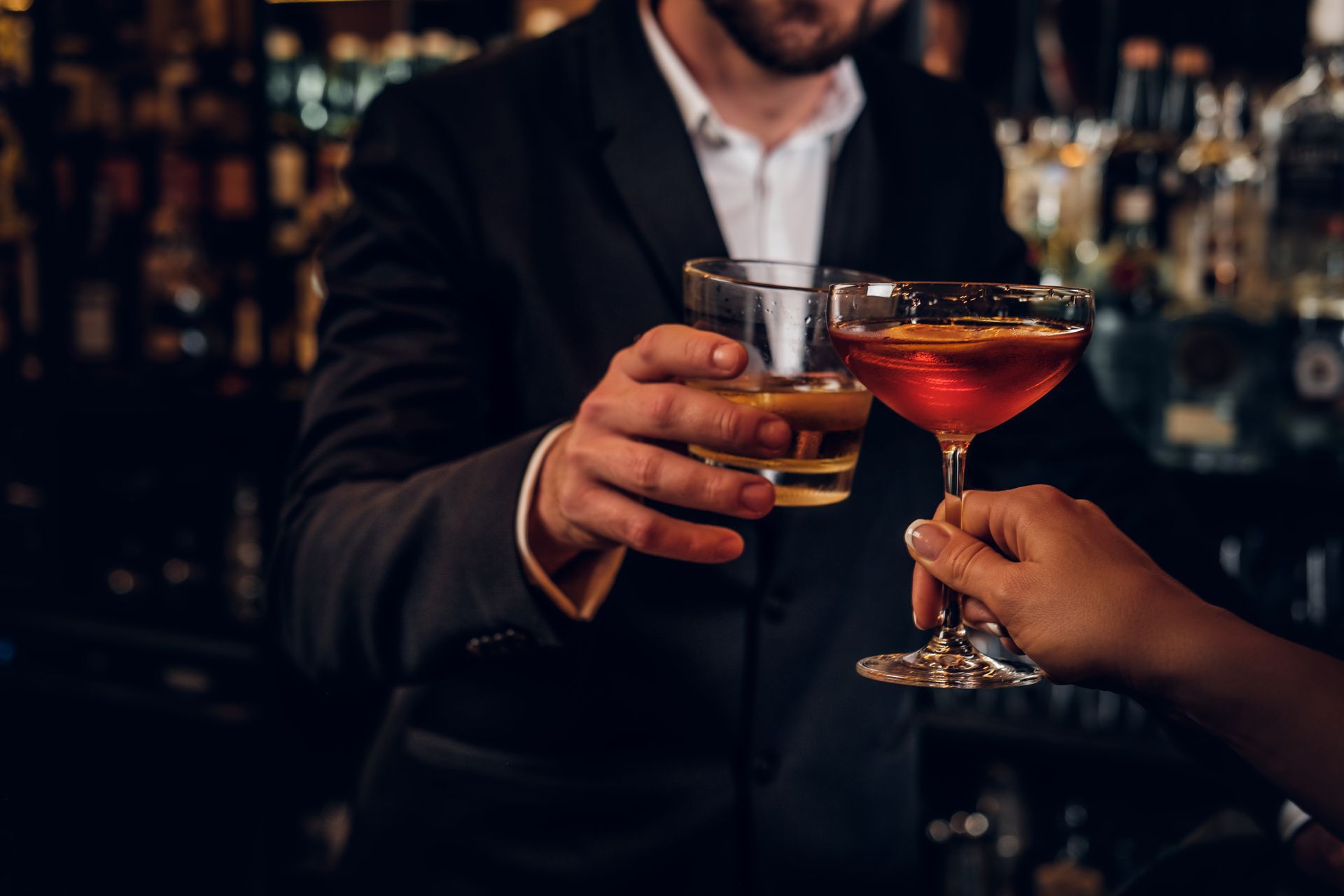 Ein Barkeeper im schwarzen Anzug prostet mit einem Cocktail auf die Hand einer Person, die ein Glas roten Cocktail hält. Barszene.
