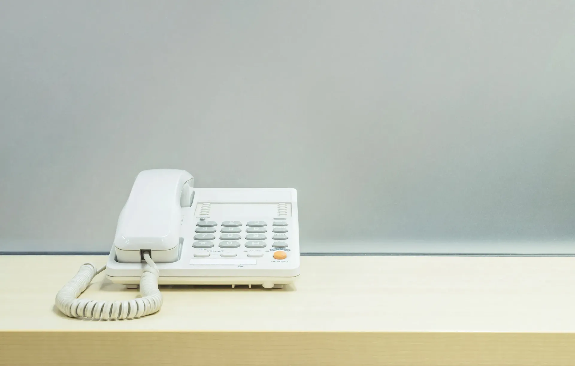 Teléfono fijo blanco sobre un escritorio de madera clara contra una pared gris.