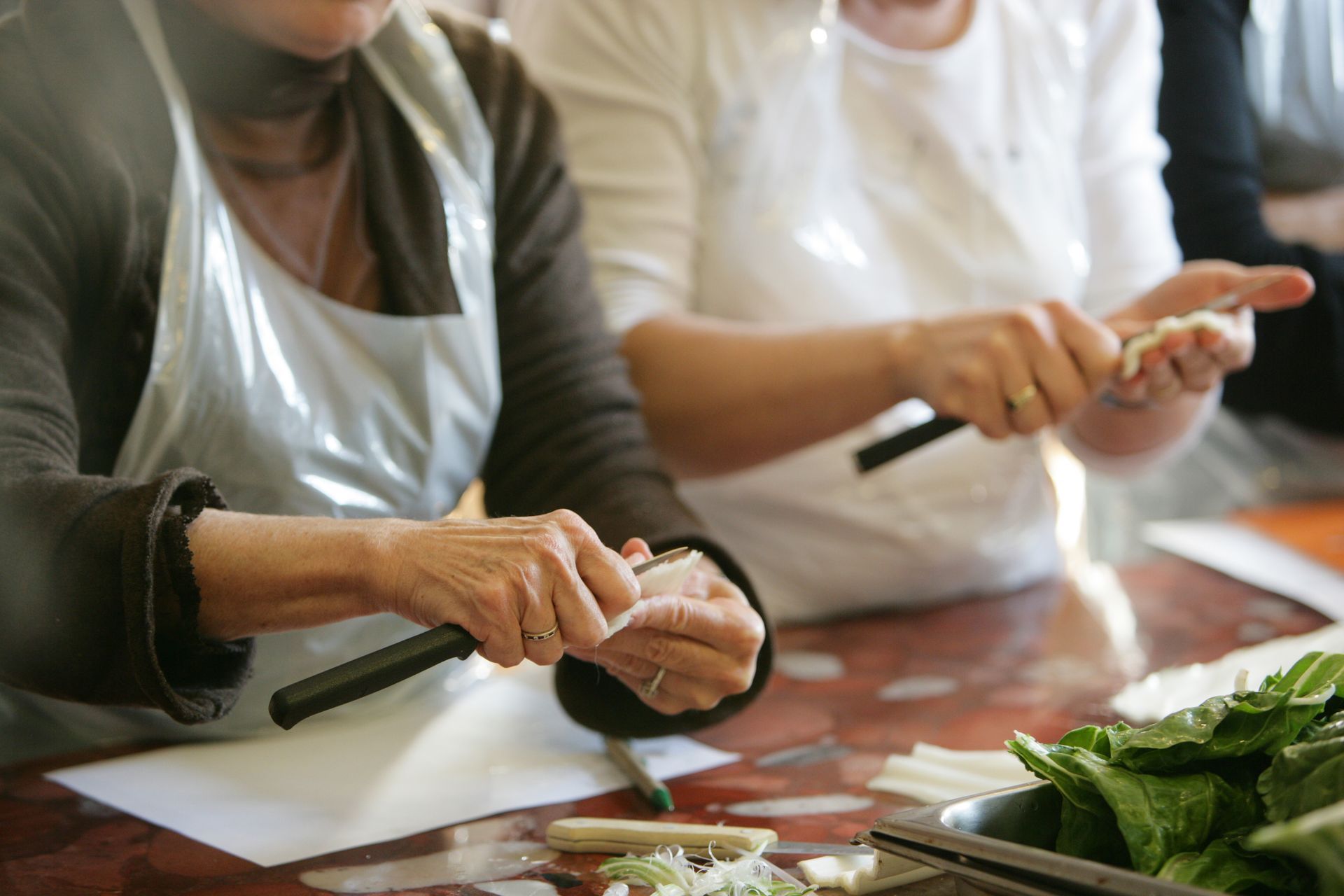 Des personnes découpent des ingrédients sur un plan de travail de cuisine, avec des légumes verts à feuilles au premier plan.