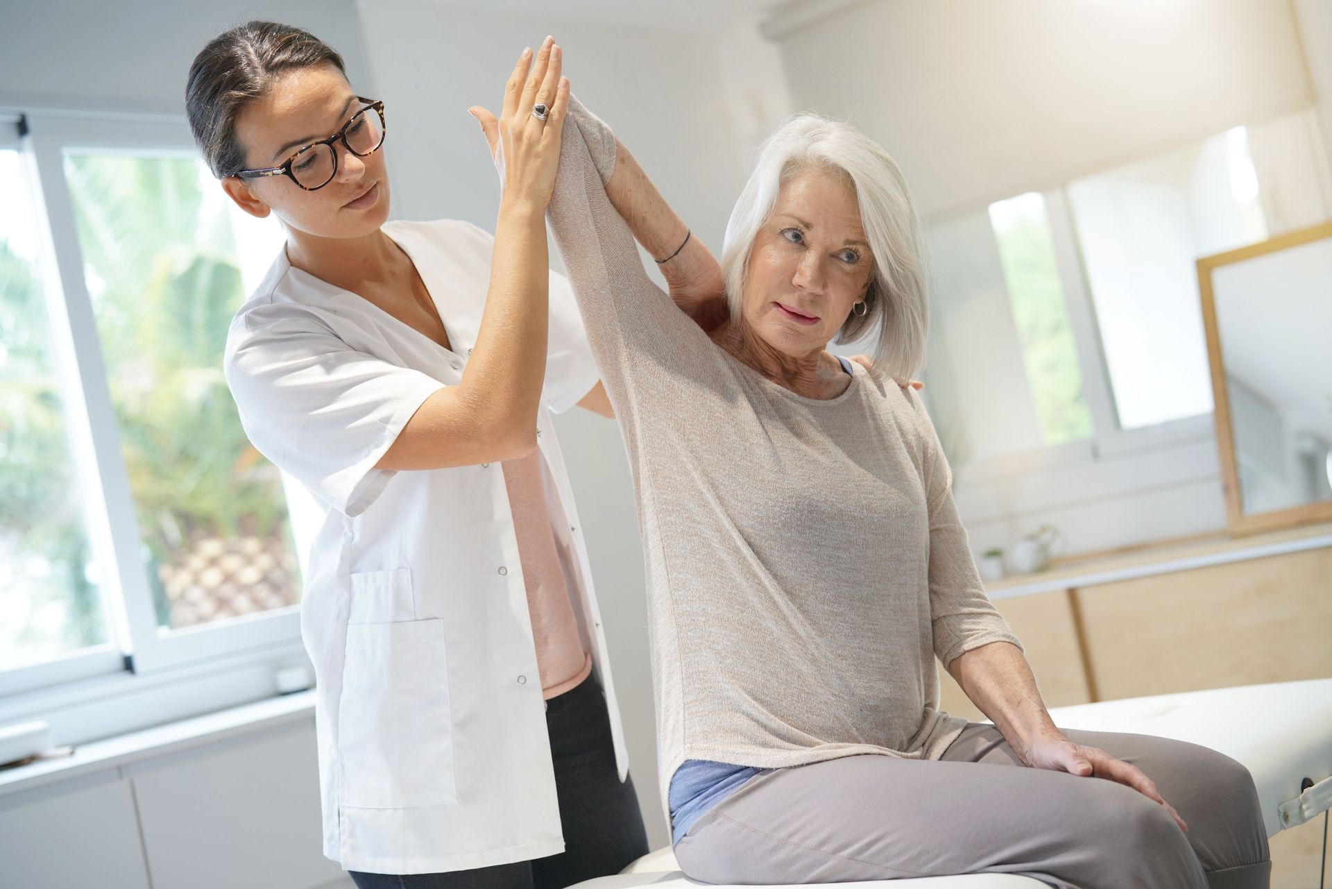 Une thérapeute aide une dame assise à effectuer un étirement des bras dans une salle lumineuse.