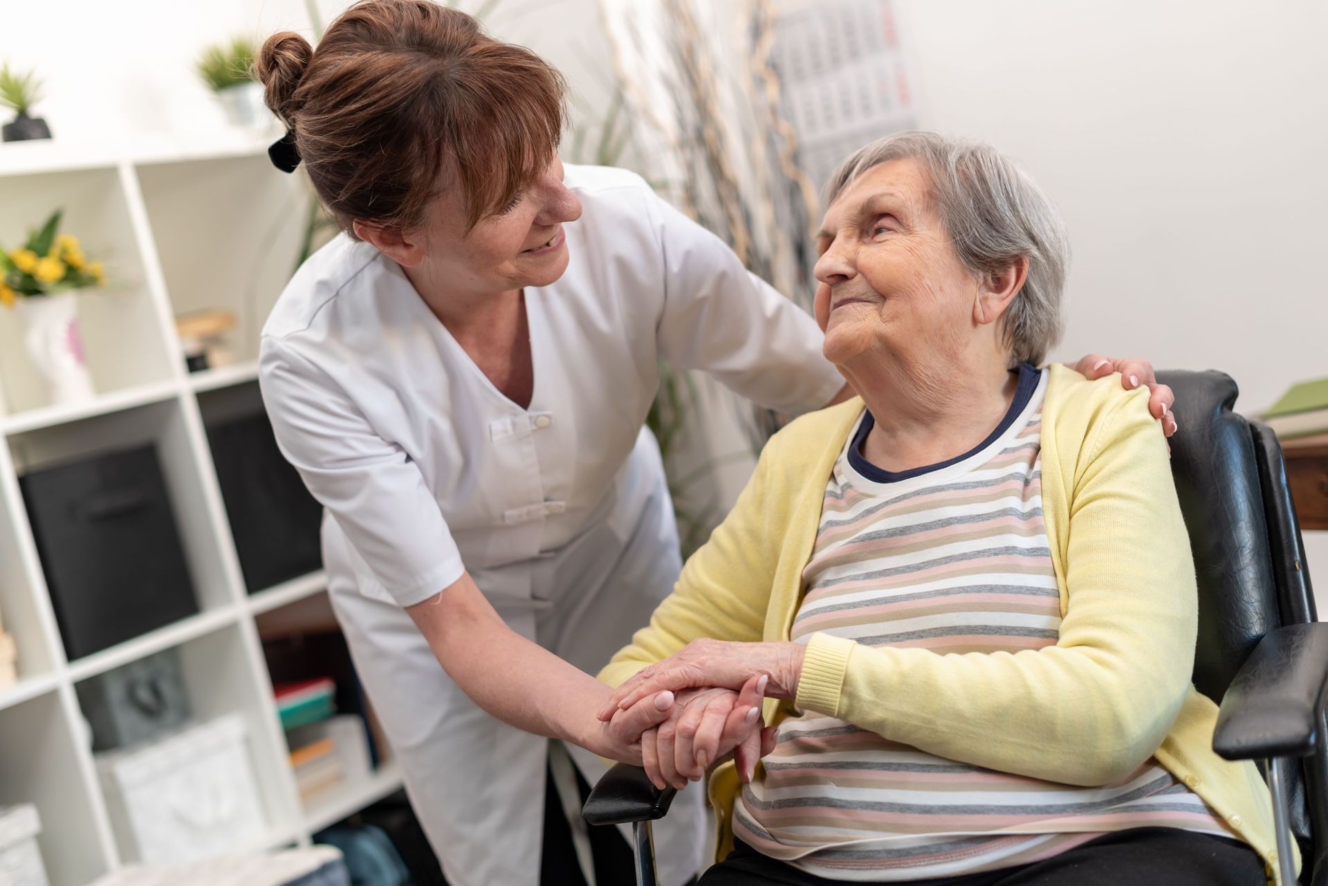 Une soignante sourit et tient la main d'un patient assis dans un cadre domestique lumineux.
