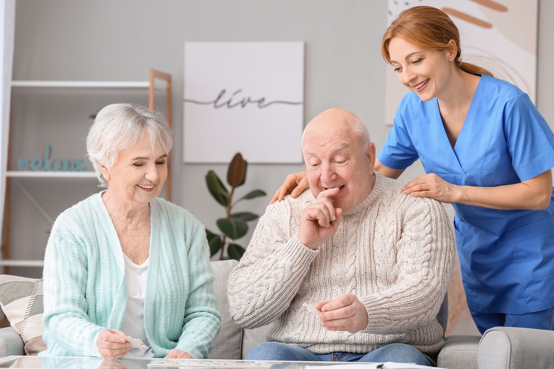 Une aide-soignante assiste un couple de personnes âgées souriantes dans un salon lumineux.