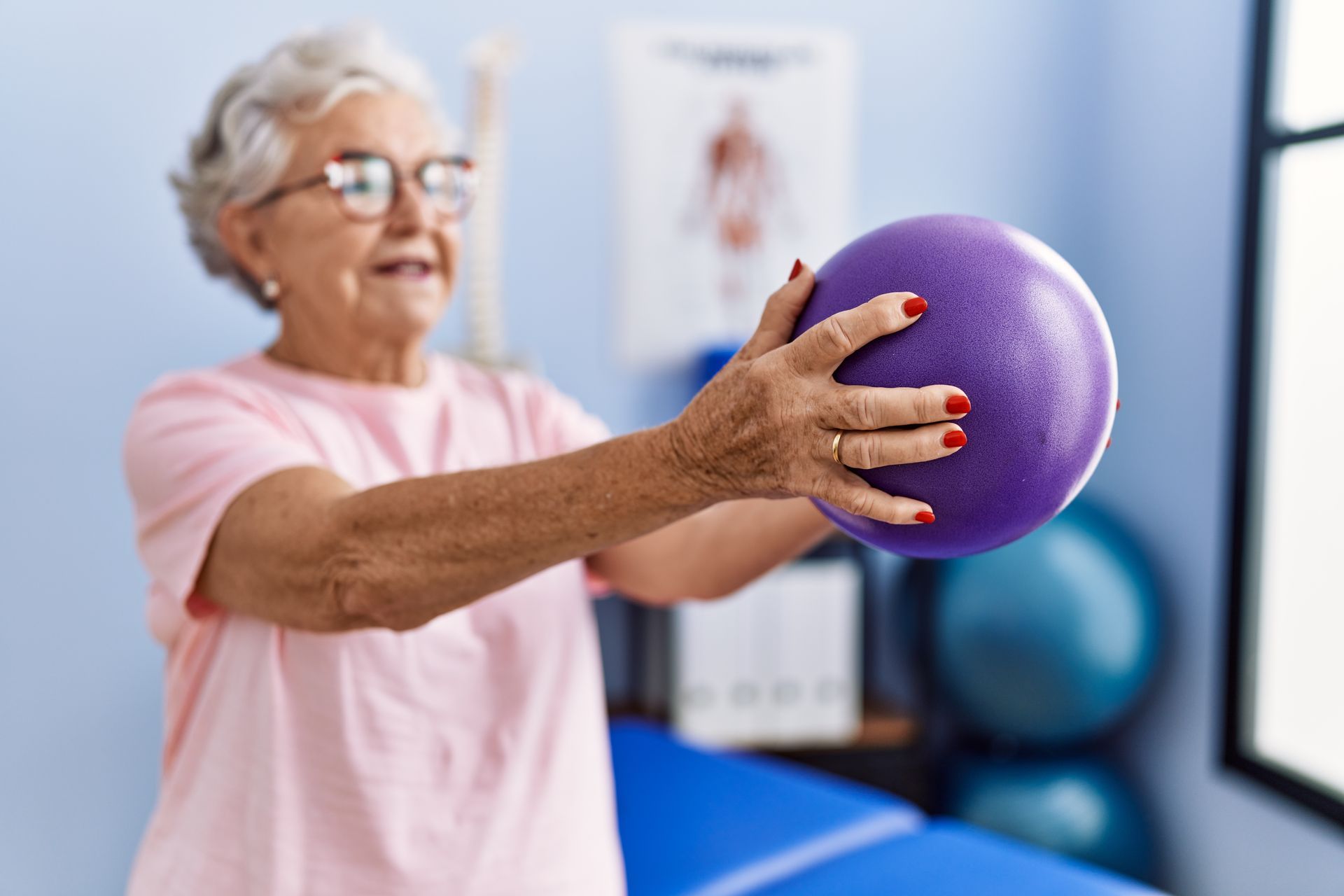 Une femme âgée en chemise rose fait de l'exercice avec un ballon violet dans une salle.